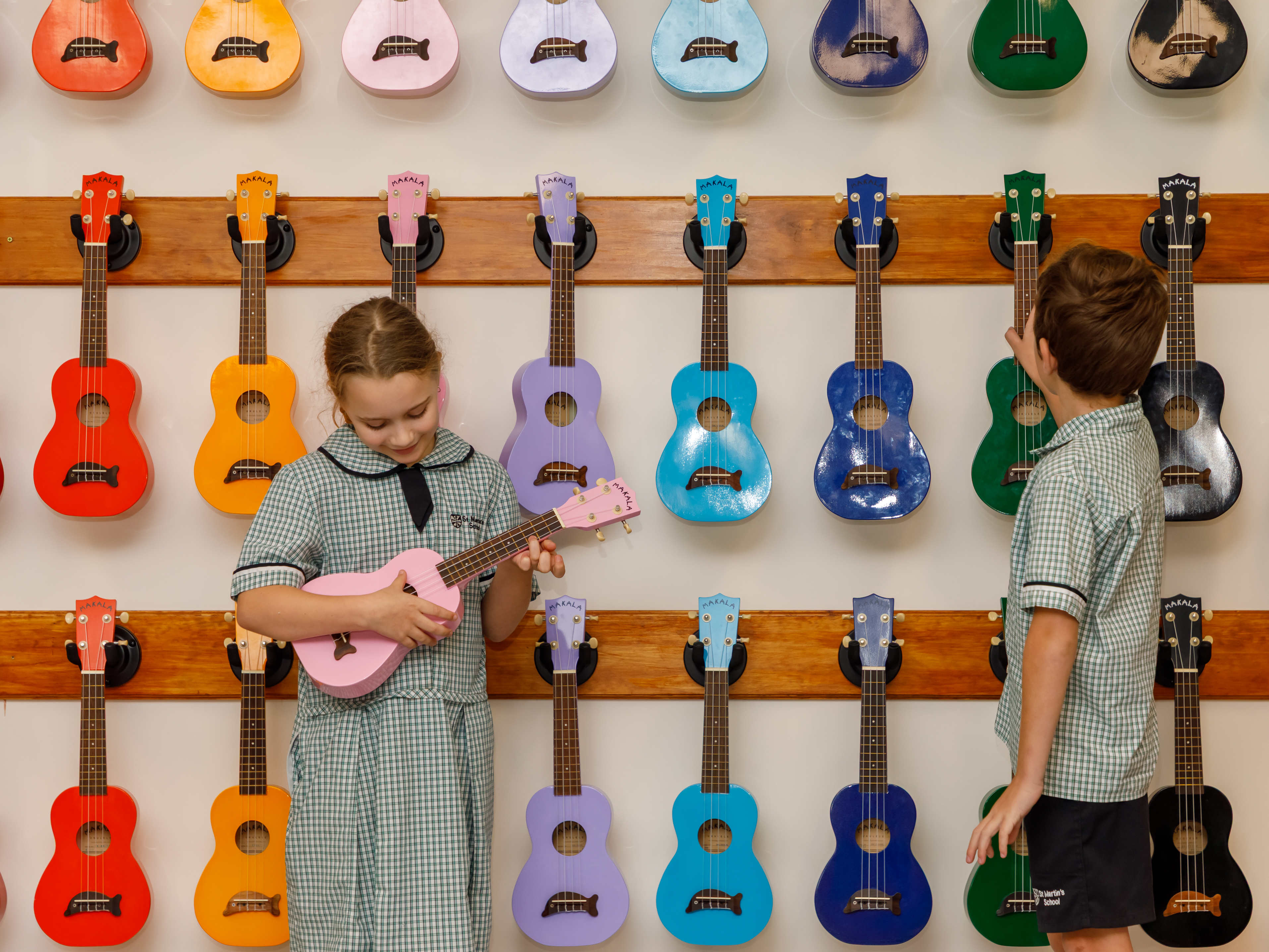 Two students are standing in front of a wall of ukeleles. 